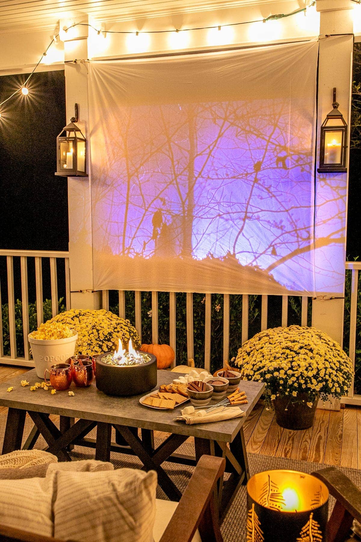 fall movie playing on an outdoor movie screen on a back porch decorated for fall with pumpkins, mums, and tabletop fire pit