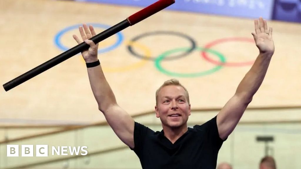 Chris Hoy, wearing a black t-shirt, holds a baton in the air during a ceremony at a velodrome in Paris. Behind him is the velodrome with the five Olympic rings